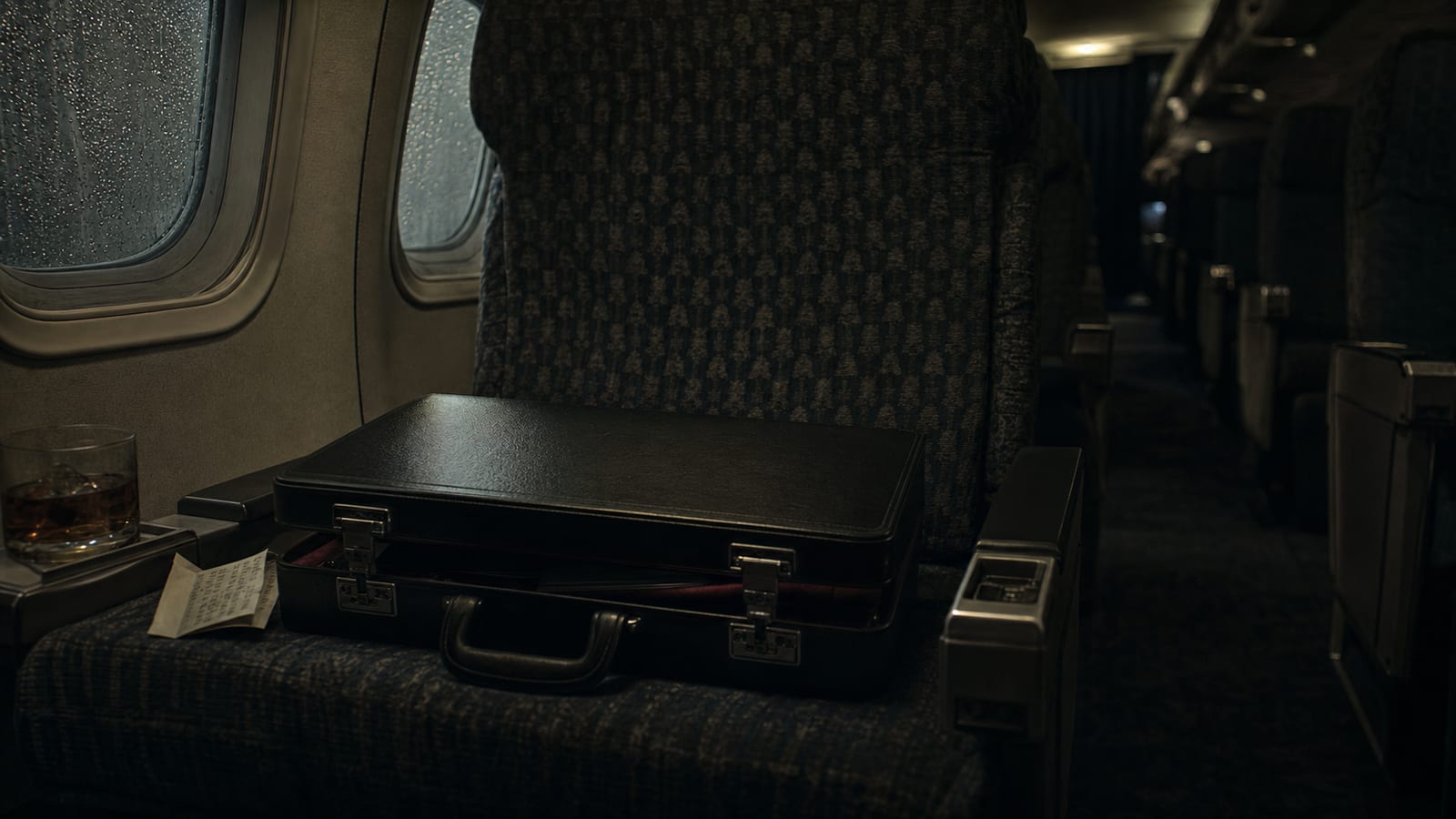 Closed attache case on a dim vintage airplane seat beside a drink and folded note