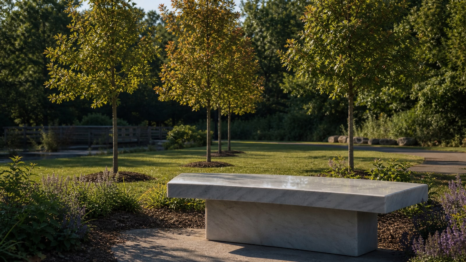 Four young oak trees and a marble bench in a quiet park, representing the memorial for Jayne Friedt, Ruth Shelton, Daniel Davis, and Mark Flemmonds.