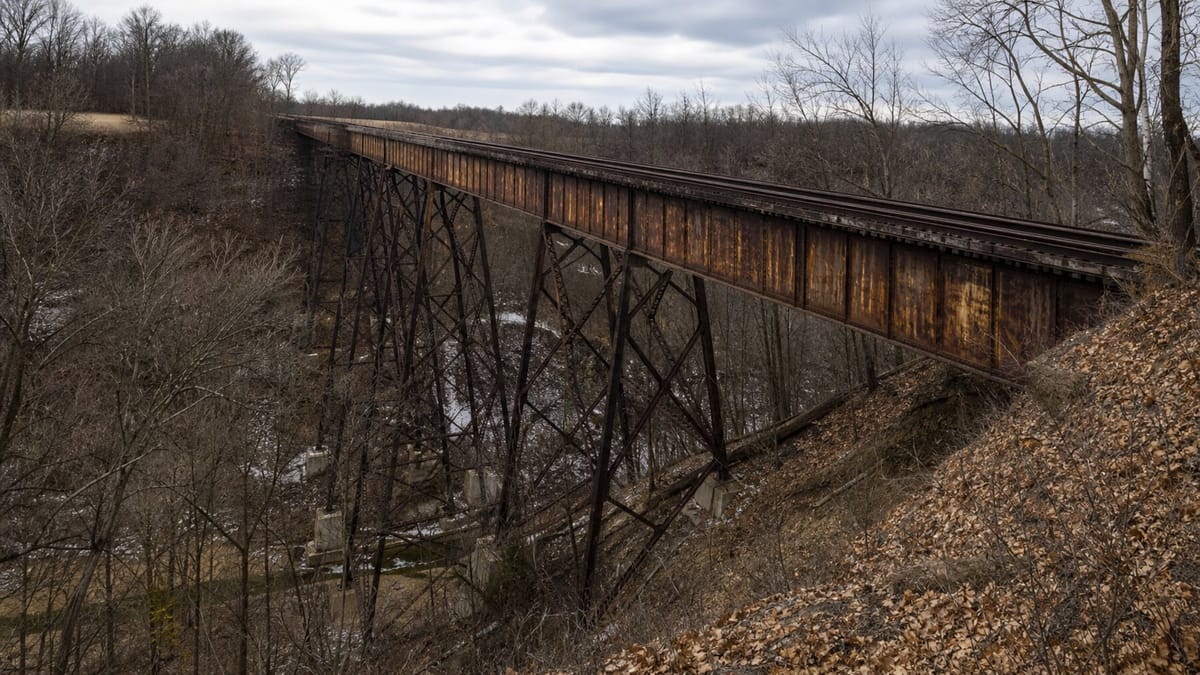 A rusted abandoned railroad trestle over late-winter Indiana woods, representing the Monon High Bridge in the Delphi murders case.