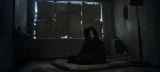 Eli sits on a bare mattress in a dark apartment room with cardboard sheets taped over the window.