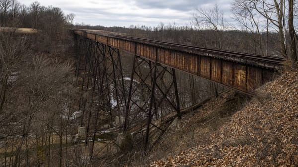 A rusted abandoned railroad trestle over late-winter Indiana woods, representing the Monon High Bridge in the Delphi murders case.